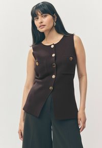 Top marrone senza maniche con una finitura texturizzata, dotato di due tasche frontali, bottoni dorati e uno spacco laterale, abbinato a pantaloni scuri.