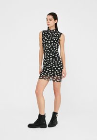 Schwarzes ärmelloses Kleid mit weißen Punkten, das ein drapiertes Design und durchscheinenden Stoff aufweist; kombiniert mit schwarzen Knöchelstiefeln.
