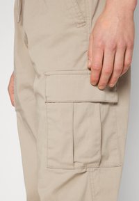 Bežové cargo kalhoty s boční kapsou na klopu, hladkou texturou látky a viditelnými švy na pásu a nohavicích.