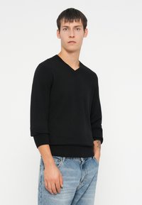 Czarny sweter z dekoltem w kształcie V z długimi rękawami, prążkowanymi mankietami i brzegiem. Model w jasnoniebieskich, postarzanych dżinsach, tatuaż widoczny na lewej dłoni.