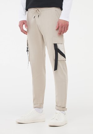 Pantaloni cargo beige con polsini elasticizzati, tasche laterali e una stampa grafica sulla tasca cargo, abbinati a sneaker bianche.