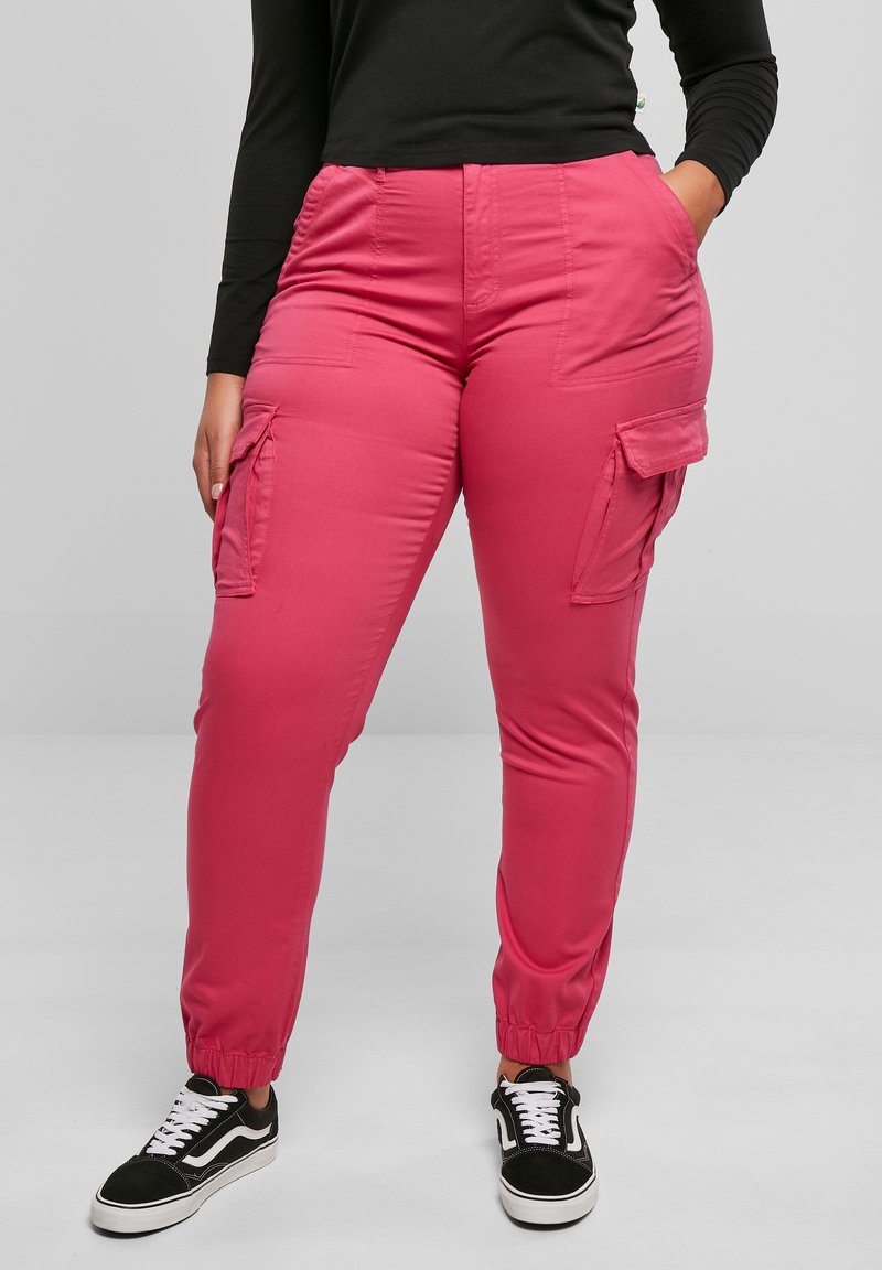 Personne portant un pantalon cargo rose vif avec des poignets élastiques, des baskets noires avec des lacets blancs et un haut noir à manches longues, debout les mains dans les poches.