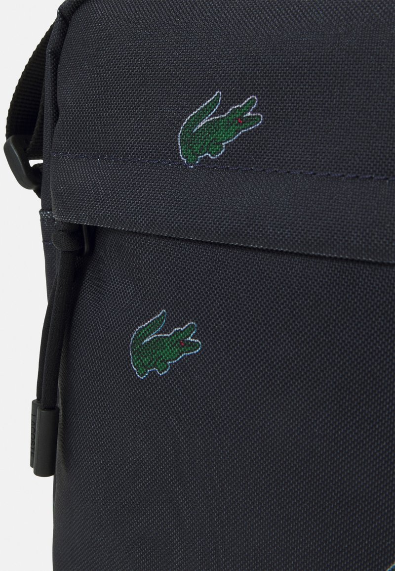 Sac en tissu noir texturé avec deux logos de crocodile brodés en vert et une sangle d'accent noir. Fermeture à zip avec quincaillerie de marque.