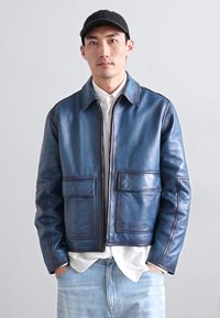 Giacca in pelle blu navy con due tasche anteriori, colletto e dettagli di cucitura visibili. Indossata sopra una camicia bianca.