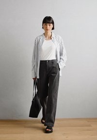 Weißes T-Shirt unter einem vertikal gestreiften, übergroßen blauen und weißen Hemd, kombiniert mit hoch taillierten grauen weiten Hosen und schwarzen Sandalen.