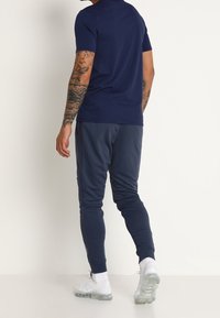 Marinblå kortärmad skjorta i kombination med mörkgrå tapered joggers med sidostråk, slät tygtextur och vita sportskor.