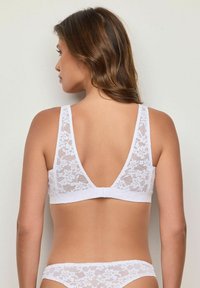 Bílá krajková braletka s V-zadním designem, zdobená květinovými vzory, širokými ramínky a háčkovým zapínáním. Měkká textura a jemné detaily.