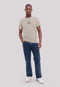 Junger Mann steht mit einer Hand in der Tasche, trägt ein beiges T-Shirt mit Grafik, blaue Jeans und weiße Sneakers vor einem hellgrauen Hintergrund.