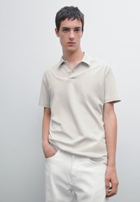 Beige polo skjorta av mjukt tyg, med kort krage, knappslå och en avslappnad passform, ihop med vita byxor.