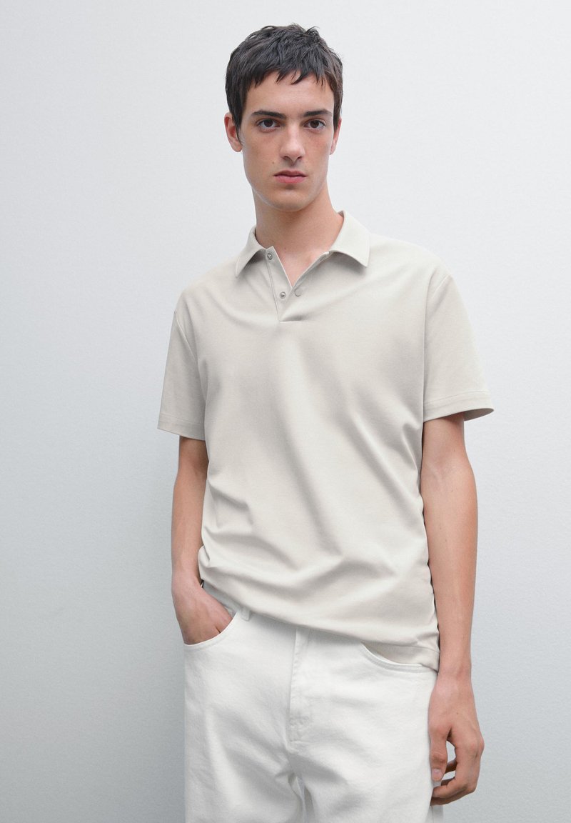 Beige polo skjorta av mjukt tyg, med kort krage, knappslå och en avslappnad passform, ihop med vita byxor.