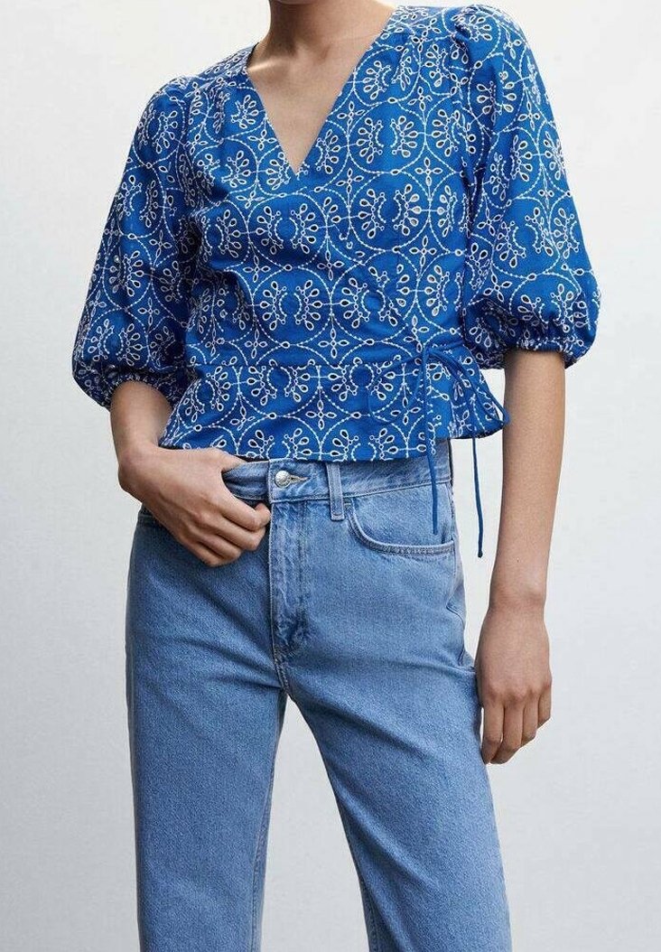 Blusa azzurra a fantasia floreale con maniche a sbuffo, scollatura a V e dettaglio con cordoncini, abbinata a jeans chiari a vita alta.