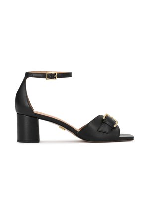 Sandale en cuir noir avec une large bride, un détail boucle et une lanière autour de la cheville. Dotée d'un talon épais pour la stabilité et d'une finition lisse.