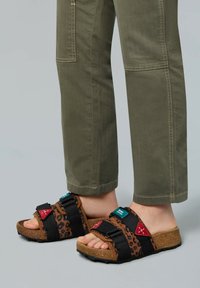Brown and black leopard print sandals with adjustable straps, cork soles, and red and teal accents, paired with olive green pants.