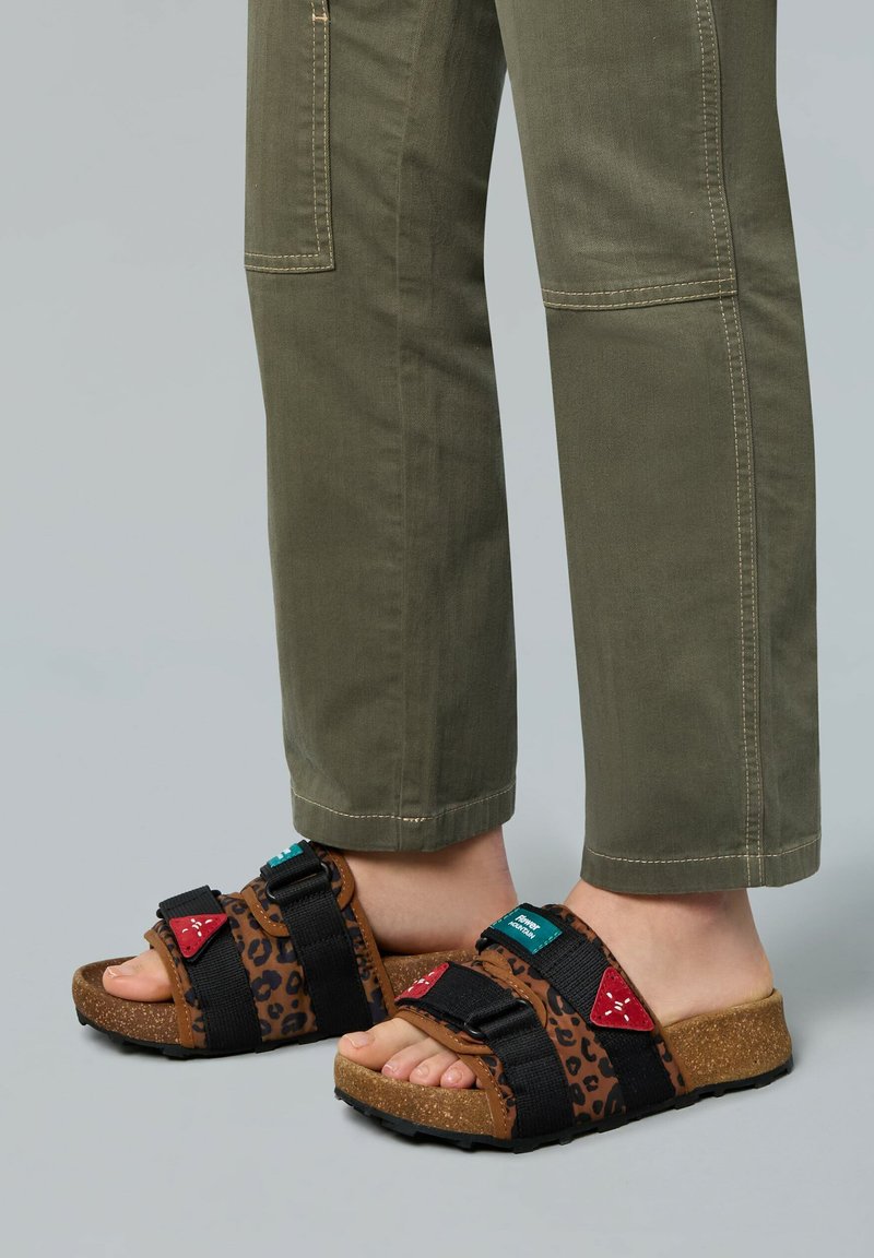 Brown and black leopard print sandals with adjustable straps, cork soles, and red and teal accents, paired with olive green pants.
