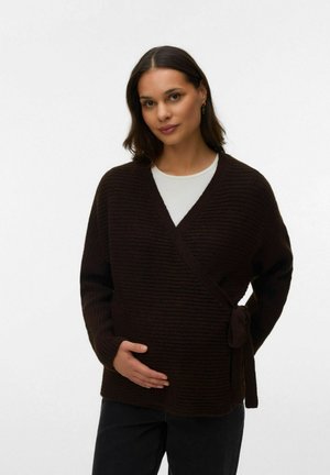 Kvinde med mørkt hår iført en brun omslagsstriktrøje over en hvid top, blidt holdende om sin mave, stående foran en ensartet lys baggrund.