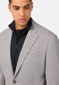Svetlo siv blazer z teksturirano površino in izrezanimi lapelami, položen čez navy zip-up srajco, ki ima ohlapen kroj in en gumb.