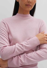 Helles rosa Langarmoberteil mit hohem Kragen. Weicher, gerippter Stoff an den Ärmeln. Das Modell schlägt die Arme vor der Brust über Kreuz und zeigt das taillierte Design des Kleidungsstücks.