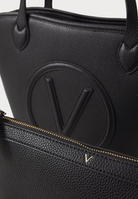 Le sac fourre-tout en cuir noir présente un extérieur texturé, un logo "V" embossé et une pochette zippée dorée avec un subtil accent "V".
