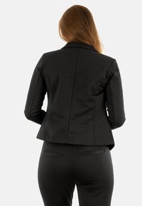 Blazer noir ajusté avec un motif à pois subtil, présentant une silhouette cintrée, des revers crantés et une texture lisse.