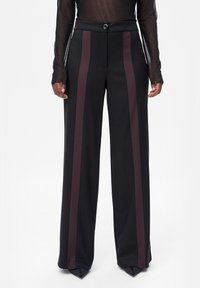 Czarne szerokie spodnie z ciemnobrązowymi pionowymi paskami, noszone z czarnymi szpiczastymi butami i przezroczystą bluzką z długim rękawem.