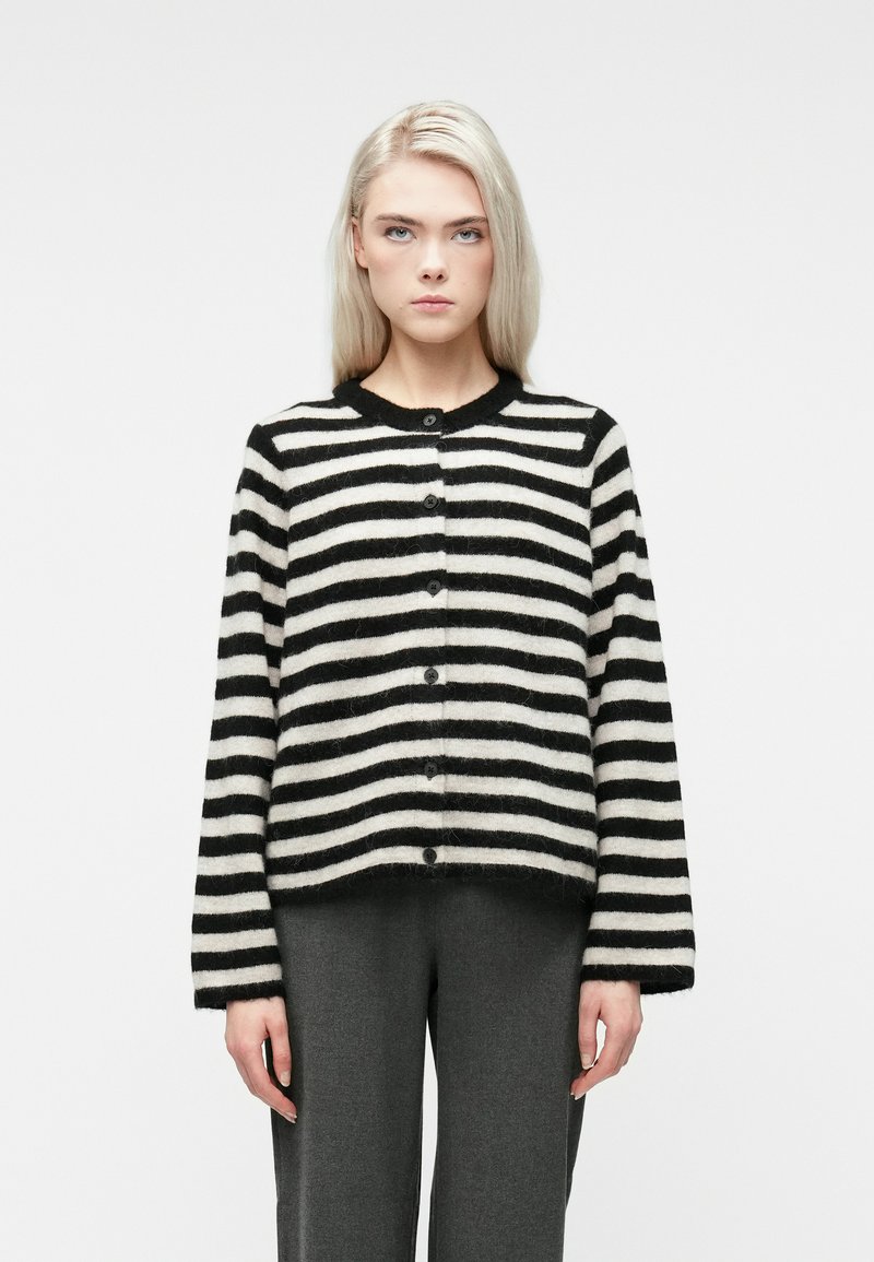 Blondynka w czarno-białej poziomej paskowanej koszuli z guzikami i ciemnoszarych spodniach, stojąca na białym tle.