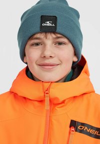 Žiarivo oranžová zip-up bunda s čiernym a oranžovým logom, vrstvená nad tealovou čiapkou s čiernym štítkom. Hladká textúra, ležérny dizajn.