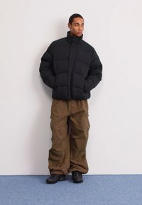 Jachetă puf din negru cu guler înalt, asortată cu pantaloni cargo maro, largi. Textură groasă, cu o culoare solidă. Booti negri completează ținuta.