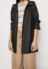 Czarna wodoodporna kurtka przeciwdeszczowa z kapturem, zatrzaskami i bocznymi kieszeniami, w połączeniu z pasiastym krótkim topem i beżowymi spodniami. Gładka faktura.