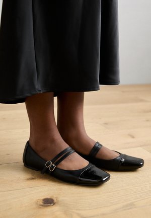 Bailarinas de charol negro con punta cuadrada, que cuentan con dos tiras con hebillas. Las suelas son planas sobre un suelo de madera clara.
