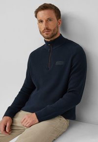 Marinefarbener Reißverschluss-Pullover mit strukturiertem Strickmuster, hohem Kragen und dezentem Logo-Detail auf der Brust, kombiniert mit hellbeigen Hosen.