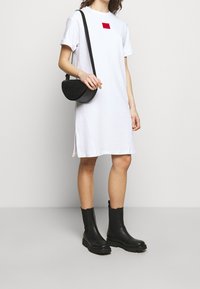 Biała sukienka do kolan z krótkim rękawem i czerwonym kwadratowym logo na piersi. Noszona z czarną torbą crossbody i czarnymi botkami za kostkę.
