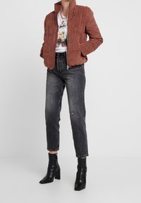 Hnědá kordurová bunda s vysokým límcem, doplněná o oprané tmavé džíny a černé kotníčkové boty na podpatku. Pod tím je jednoduché bílé tričko s grafikou.