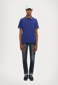 Námornícky polo tričko s vzorovaným golierom a okrajmi rukávov, s vreckom na ľavej strane, skombinované s tmavými džínami a čiernymi teniskami. Ľahká čiapka na hlave.