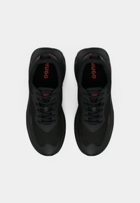 Sorte atletiske sko med et stilrent design; laget av teksturert materiale. Har svarte lisser og rød "HUGO"-logo på tungen og siden.