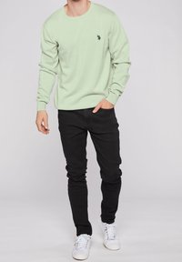 Lichtgroen gebreid sweater met een ronde hals, gecombineerd met zwarte slim-fit jeans en witte sneakers. Heeft een klein donker logo op de borst.