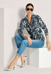 Frau in einem gemusterten Blazer, blauen, hochgekrempelten Jeans, silbernen High Heels, großen Armbändern und Sonnenbrille, sitzt auf einem weißen Block in einem minimalistischen Umfeld.