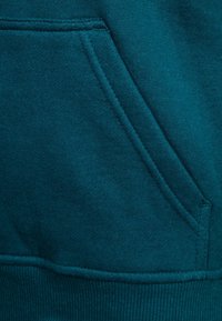 Felpa color teal in tessuto morbido con tasca anteriore e fondo a costine. I punti di cucitura sono visibili lungo i bordi.