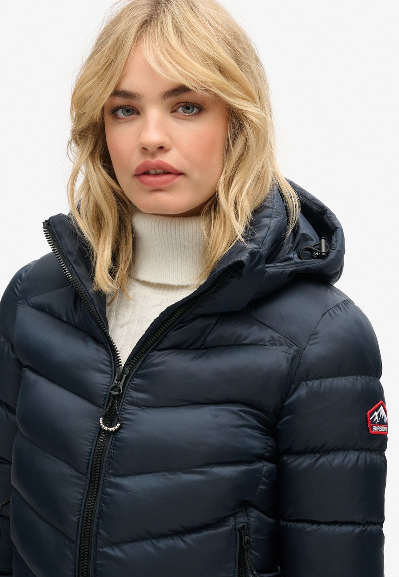 Marineblauwe gewatteerde jack met capuchon, voorzien van een rits aan de voorkant, een geborduurd berglogo op de schouder en een gekwalificeerd textuur.