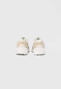 Bežas un bēšas krāsas sporta apavu pāris, redzams no aizmugures uz vienkārša baltā fona, ar sieta un zamša detaļām.