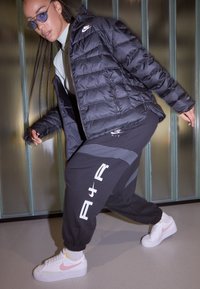 Giacca imbottita nera con design trapuntato, pantaloni della tuta neri abbinati con grafiche bianche e sneakers bianche con accenti rosa.