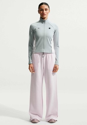 Giovane donna in piedi che indossa una giacca grigio chiaro con zip, pantaloni larghi rosa pallido e sneaker abbinate su uno sfondo bianco semplice.