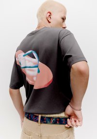 Grå kortærmet T-shirt med et stort grafisk design på ryggen i pink, blå og hvid. Bælte med blå denimtekstur og røde accenter.