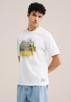 Junger Mann mit lockigem Haar, der ein weißes Grafik-T-Shirt mit Tennisplatz- und Haus-Illustration trägt, kombiniert mit hellblauen Jeans.