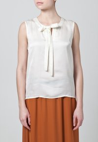 Krémová bezrukávová blůza s detaily mašle u výstřihu, vyrobená z hladkého materiálu, doplněná sukní v rezavém odstínu.