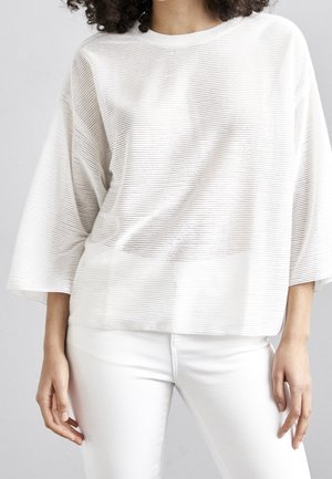 Femme portant un top blanc ample avec des lignes texturées horizontales et un pantalon blanc ajusté, debout devant un fond uni.