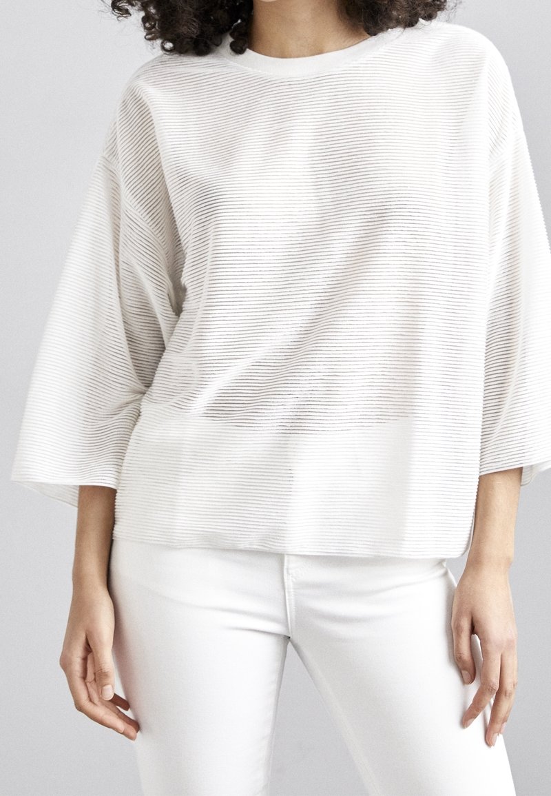 Femme portant un top blanc ample avec des lignes texturées horizontales et un pantalon blanc ajusté, debout devant un fond uni.