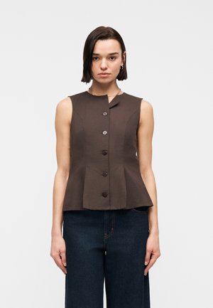 Kvinne med kort mørkt hår iført ermeløs mørkebrun knappet topp og mørkeblå jeans, stående mot en ensfarget hvit bakgrunn.