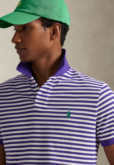 Άνδρας που φοράει πράσινο καπέλο και πουκάμισο πόλο με μωβ και άσπρες ρίγες, με σηκωμένο γιακά, κοιτάζοντας προς τα αριστερά μπροστά σε απλό φόντο.