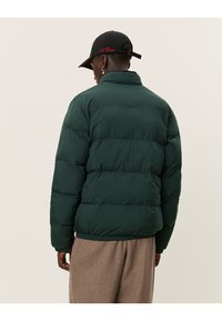 Green puffer jacket with a boxy design, featuring horizontal stitching and an elastic hem. The back has a high collar and a matte finish.