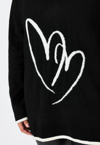 Schwarzer Pullover mit weichem Stoff, großem weißen Herzdesign und kontrastierendem weißen Saum und an den Bündchen.